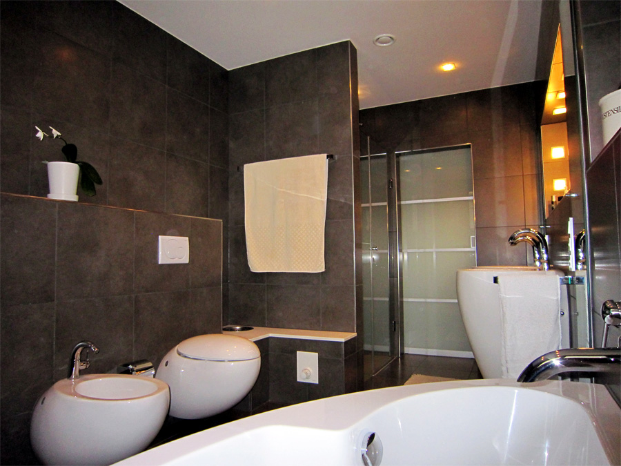 Modernes Badezimmer mit großformatigen Anthrazitfliesen, wandhängendem Bidet und Toilette, Trockenbauelementen, einer Badewanne, einem Doppelwaschbecken und einer bodengleichen Dusche. 