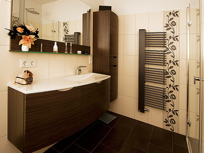 Badezimmer mit weißen Wandfliesen, schwarzen Bodenfliesen, bodengleicher Dusche, schwarzem Handtuchwärmer, weißem Waschbecken mit dunklem, wandhängendem Unterschrank.